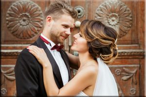 Wedding photo shooting. Bridegroom and bride standing near wooden door and embracing each other. Outdoor, waist up, closeup